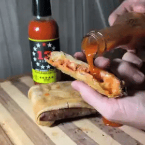 Person pouring hot sauce onto a sandwich with a bottle of hot sauce in the background.